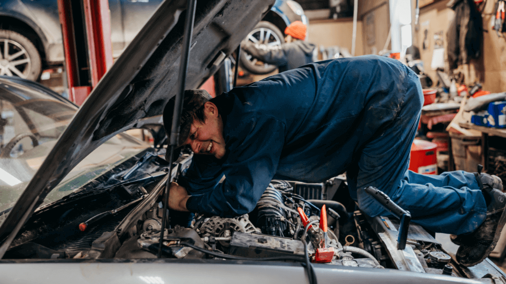 24-hour Towing | Rowes Garage Inc. in Corinth, ME. Image of a mechanic reaching under a car hood to repair an engine. In the background is another mechanic focused on another vehicle.